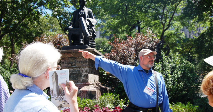 Fred stands in front of a crowd and points to a statue while giving the Flatiron Free Walking Tour in Madison Square Park. 